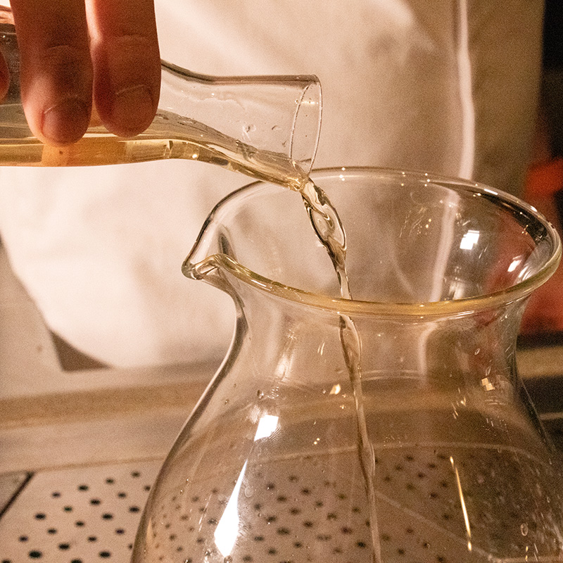Cordial being poured from a small carafe to a large glass jar