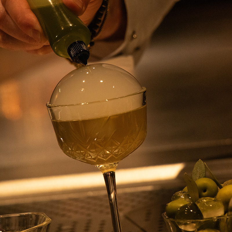 Nocellara Del Belice Olive Martini finished with a flavour bubble and being popped by a olive oil dropper