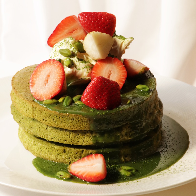 Pistachio and Matcha Pancakes stacked and topped with White Chocolate and Pistachio Sauce, whipped cream, Bronte pistachios, fresh strawberries and dusted with matcha powder