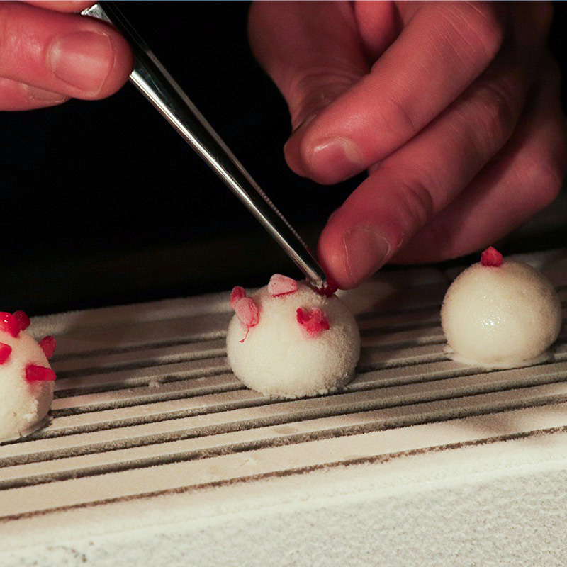 A sphere of lychee sorbet being garnished with smashed raspberries using tweezers
