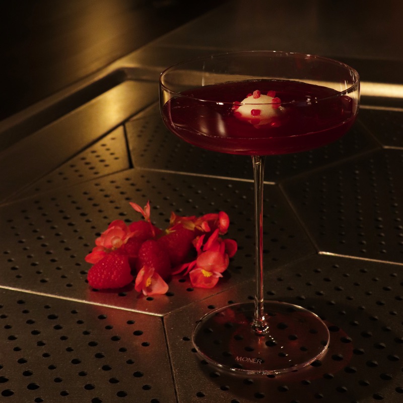 An image of the Raspberry, lychee and hibiscus "Sgroppino" Cocktail sat on a bar top with some flowers and raspberries seen just behind.