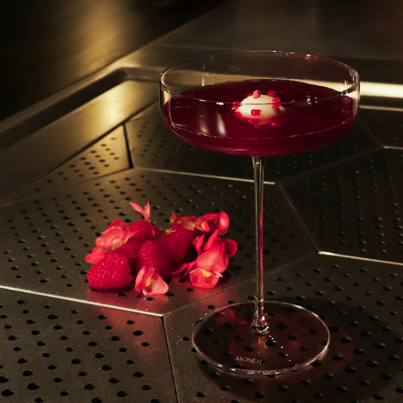 An image of the Raspberry, lychee and hibiscus "Sgroppino" Cocktail sat on a bar top with some flowers and raspberries seen just behind.